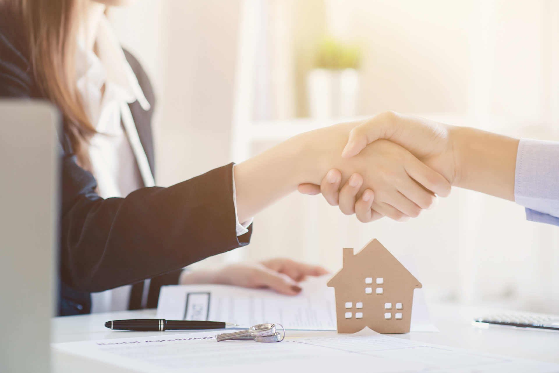 A woman shaking hands with someone while sitting at a desk with contracts, keys, a pen and a wooden cut out of a house